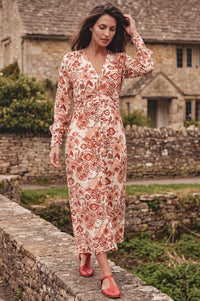 A woman wearing a long-sleeve midi dress with a rust-red and beige floral/tribal pattern, walking along a stone path in front of a rustic stone cottage.