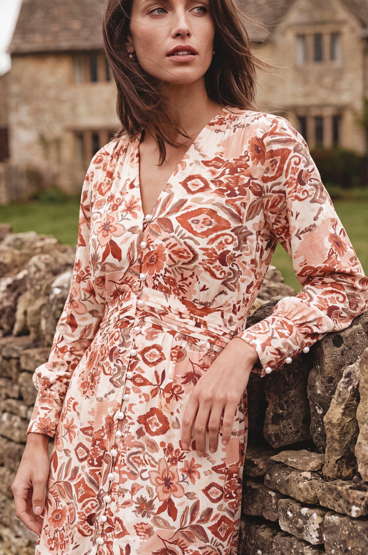 A woman wearing a long-sleeve wrap dress with a rust-orange and beige botanical pattern, leaning against a rustic stone wall with a stone building in the background.