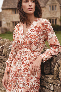 A woman wearing a long-sleeve wrap dress with a rust-orange and beige botanical pattern, leaning against a rustic stone wall with a stone building in the background.