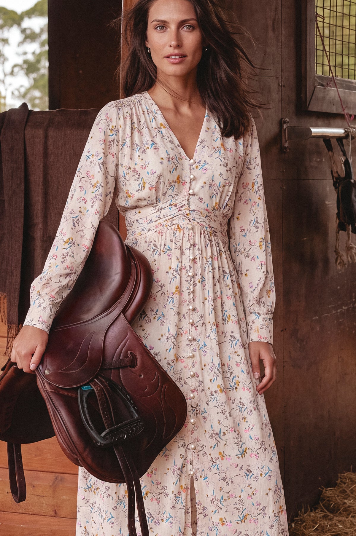 A woman stands in a rustic barn holding a dark brown leather saddle. She is wearing a long-sleeve, V-neck, midi dress with a delicate multicolor floral print on a light cream background.