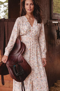 A woman stands in a rustic barn holding a dark brown leather saddle. She is wearing a long-sleeve, V-neck, midi dress with a delicate multicolor floral print on a light cream background.