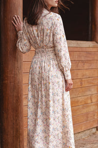 A woman wearing a long-sleeved, high-neck maxi dress with a delicate multicolor floral print on a cream background, standing next to a rustic wooden post.