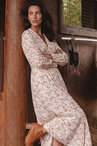 A woman wearing a long-sleeve cream maxi dress with a small multicolor floral print and tan ankle boots, leaning against a rustic wooden post in an outdoor barn setting.