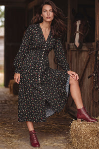 A woman stands in a rustic barn wearing a long-sleeve black midi dress with a small multicolor floral print and dark red ankle boots. A horse is visible in the background near a wooden stable door.