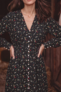 A woman wearing a long-sleeve black dress with a delicate red, white, and green botanical print, featuring a V-neckline and a button-down front.
