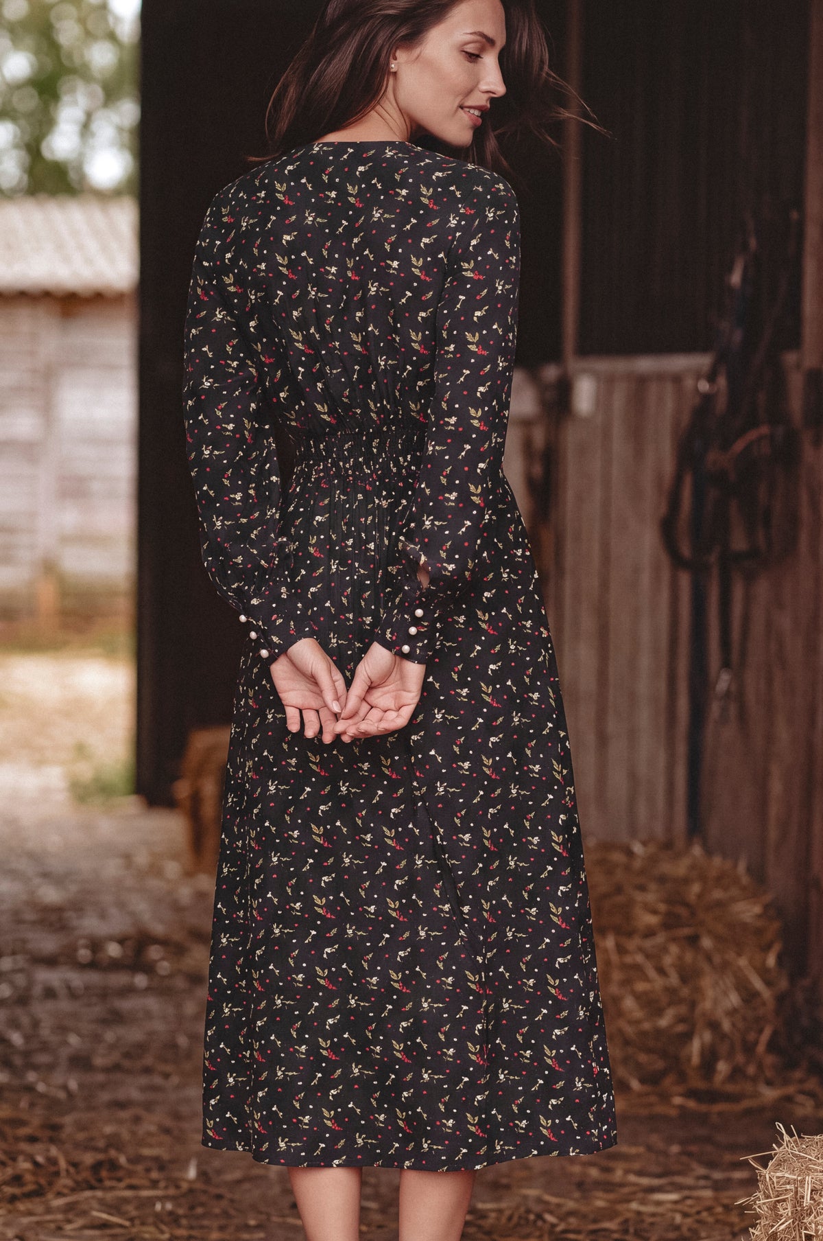 A woman wearing a long-sleeve midi dress with a small multicolor floral print on a black background, photographed from behind in a barn setting.
