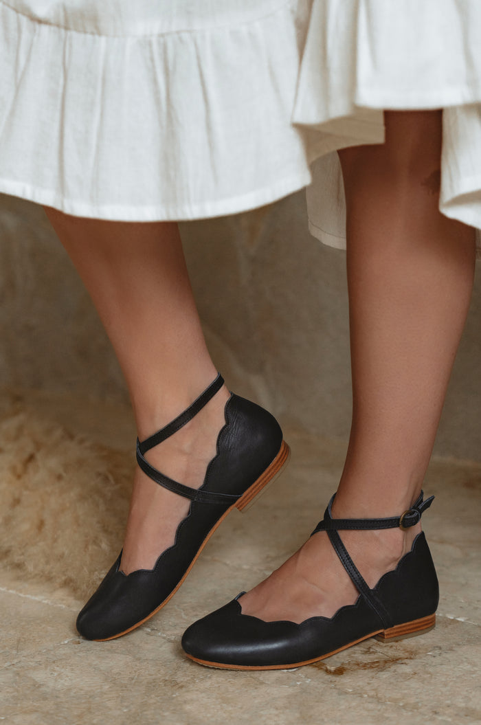 Black flat shoes with thin straps worn with a white skirt on a neutral background