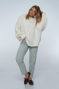 Woman wearing a white chunky knit sweater and light gray pants on a white background