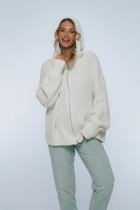 Woman wearing a white ribbed sweater and light gray pants on a white background