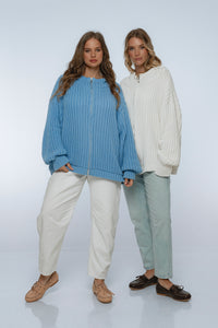Two women wearing ribbed knit sweaters and pants on a white background