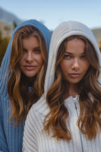 Two women wearing knit hoodies with a blurred natural background