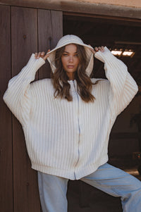 Woman wearing a white oversized sweater with a hood, standing against a wooden wall.
