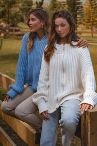 Two women sitting on a wooden bench outdoors, wearing cozy sweaters.
