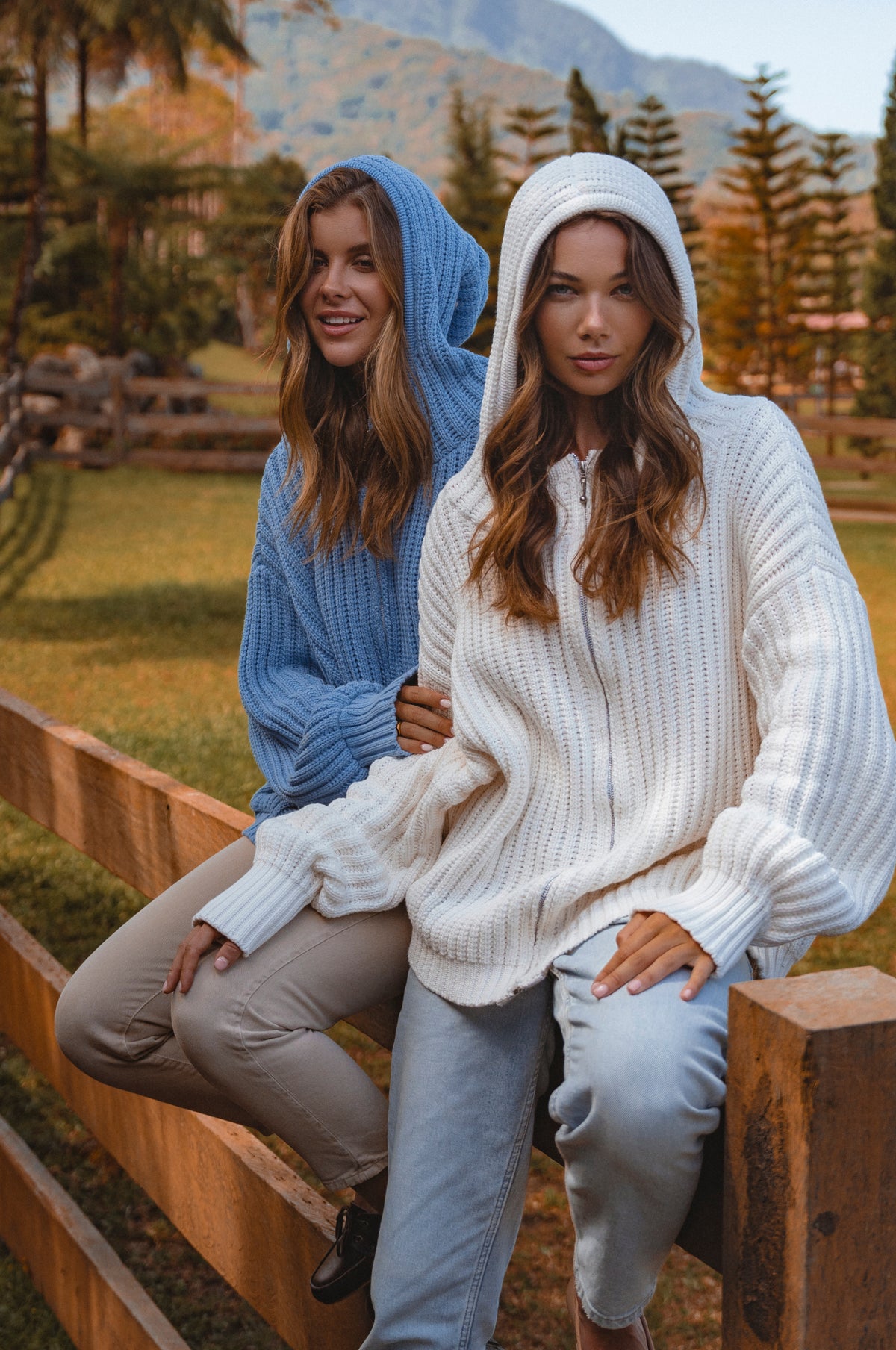 Two women wearing hoodies sitting on a wooden bench with a scenic background.