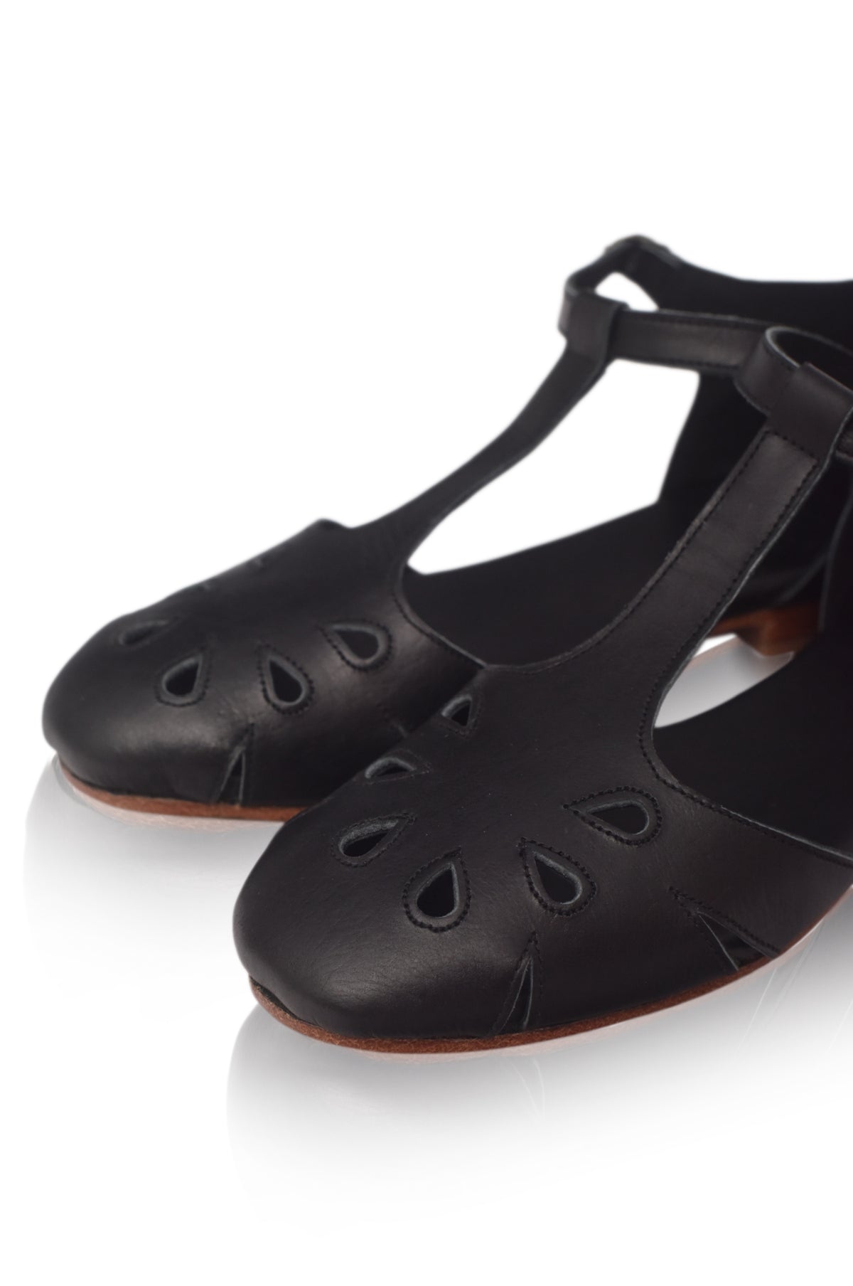 A pair of black leather T-strap ballet flats with decorative cutout details on the toe, shown on a white reflective background.