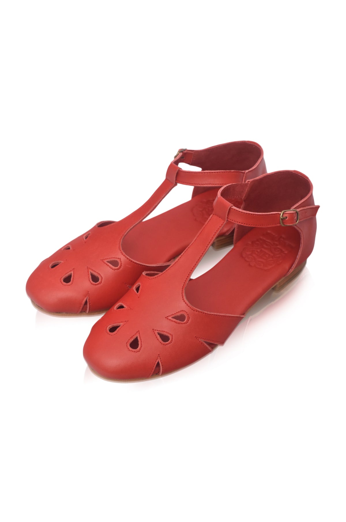 A pair of red leather T-strap flat shoes with decorative cutouts on the toe, displayed on a clean white background.
