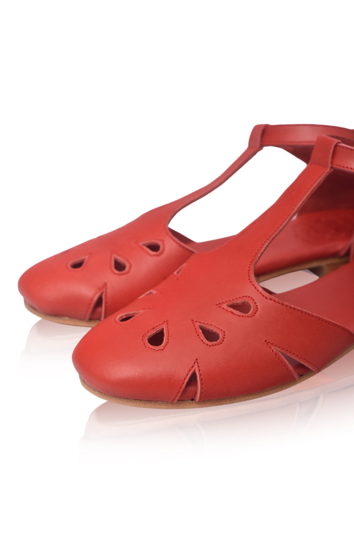 A pair of red leather flat sandals with cutout details on the toe and T-strap, shown on a reflective white surface.