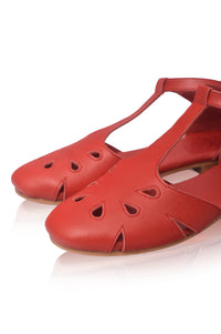 A pair of red leather flat sandals with cutout details on the toe and T-strap, shown on a reflective white surface.