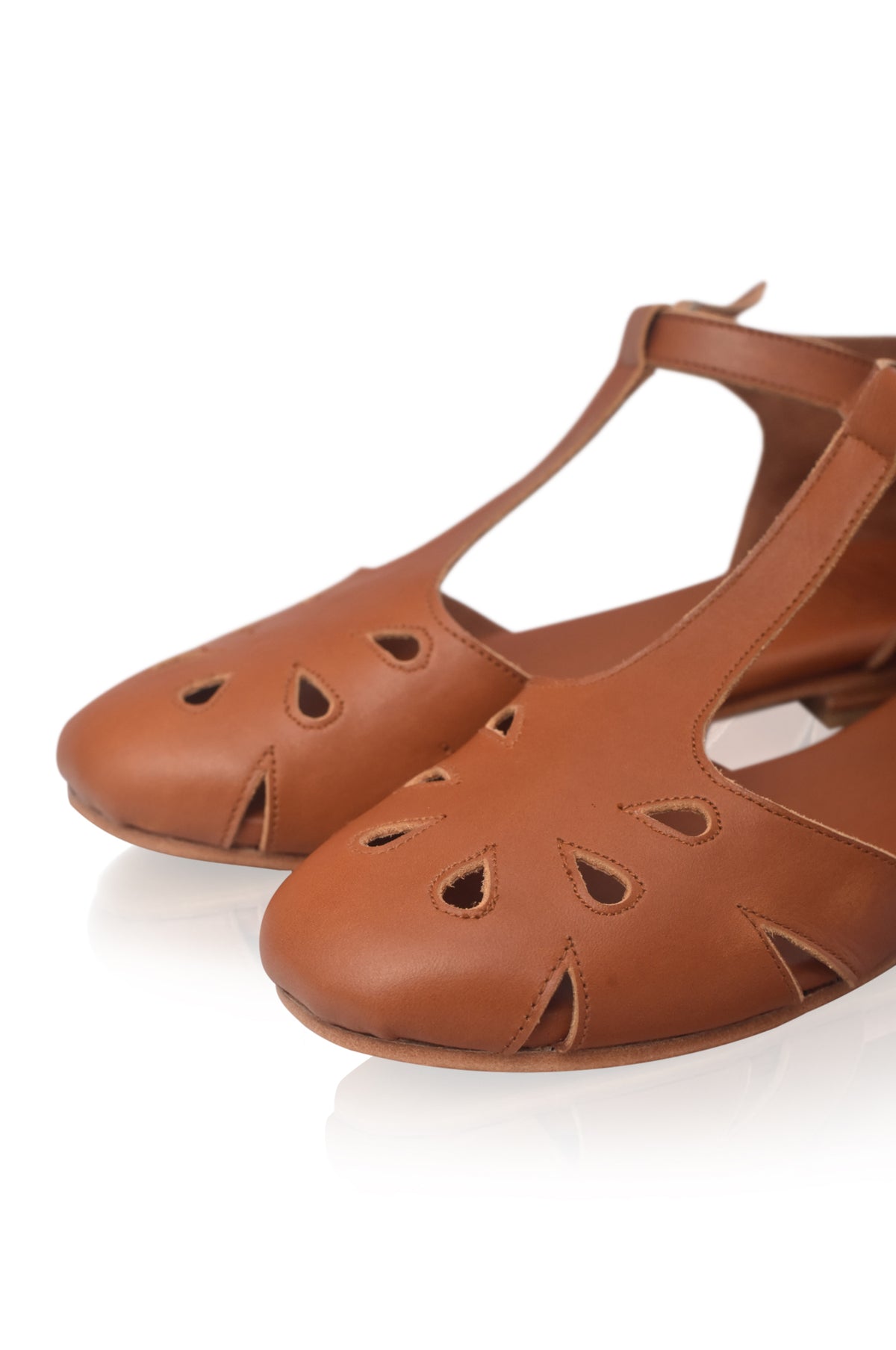 A pair of women's flat sandals in tan leather, featuring decorative cutout details on the toe and a T-strap design with an ankle strap, shown on a white reflective background.