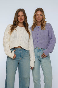 Two women wearing cable knit sweaters and jeans against a white background