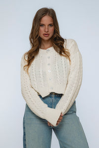 Woman wearing a white cable knit sweater and blue jeans against a plain background