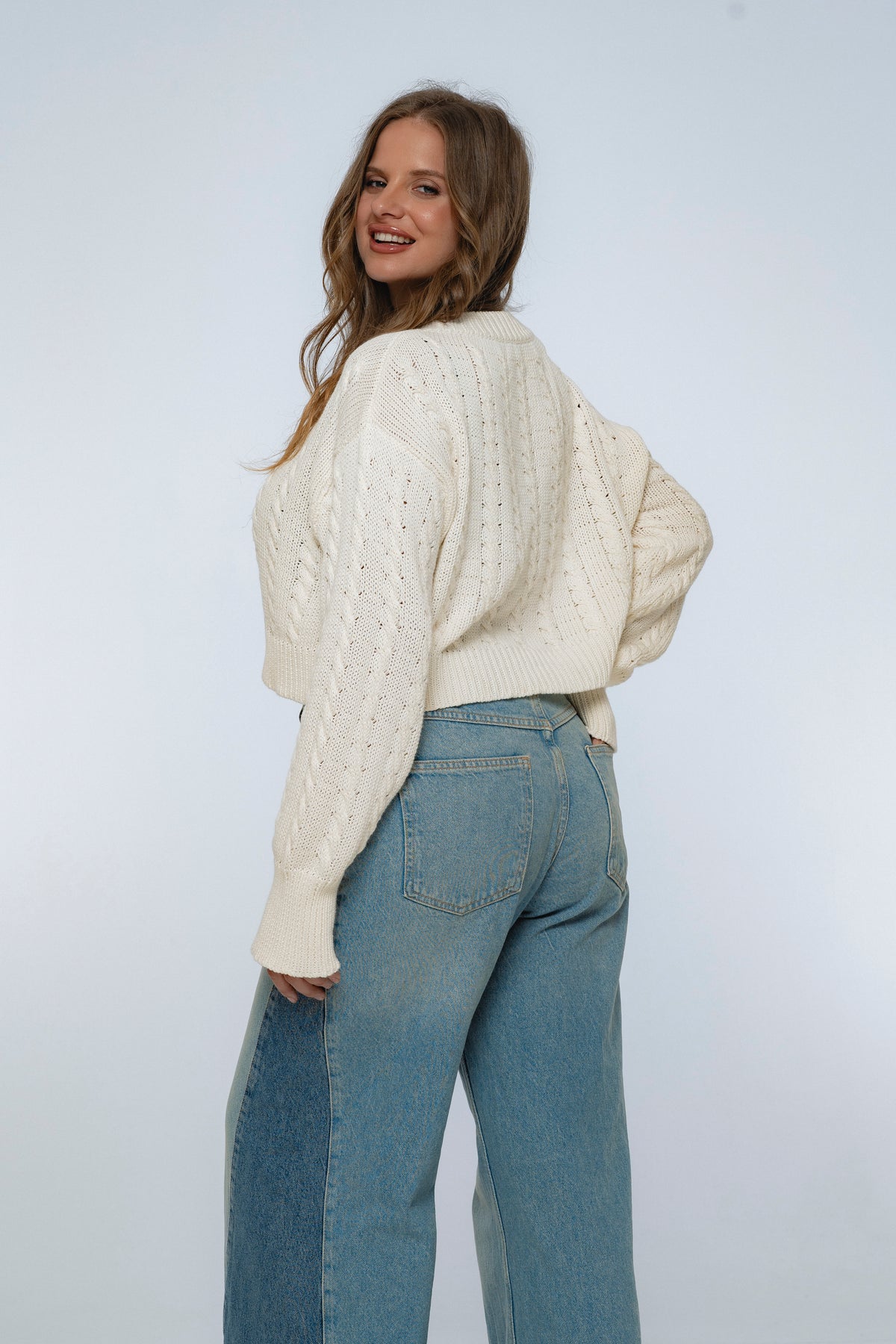 Woman wearing a cream knitted sweater and blue jeans against a white background