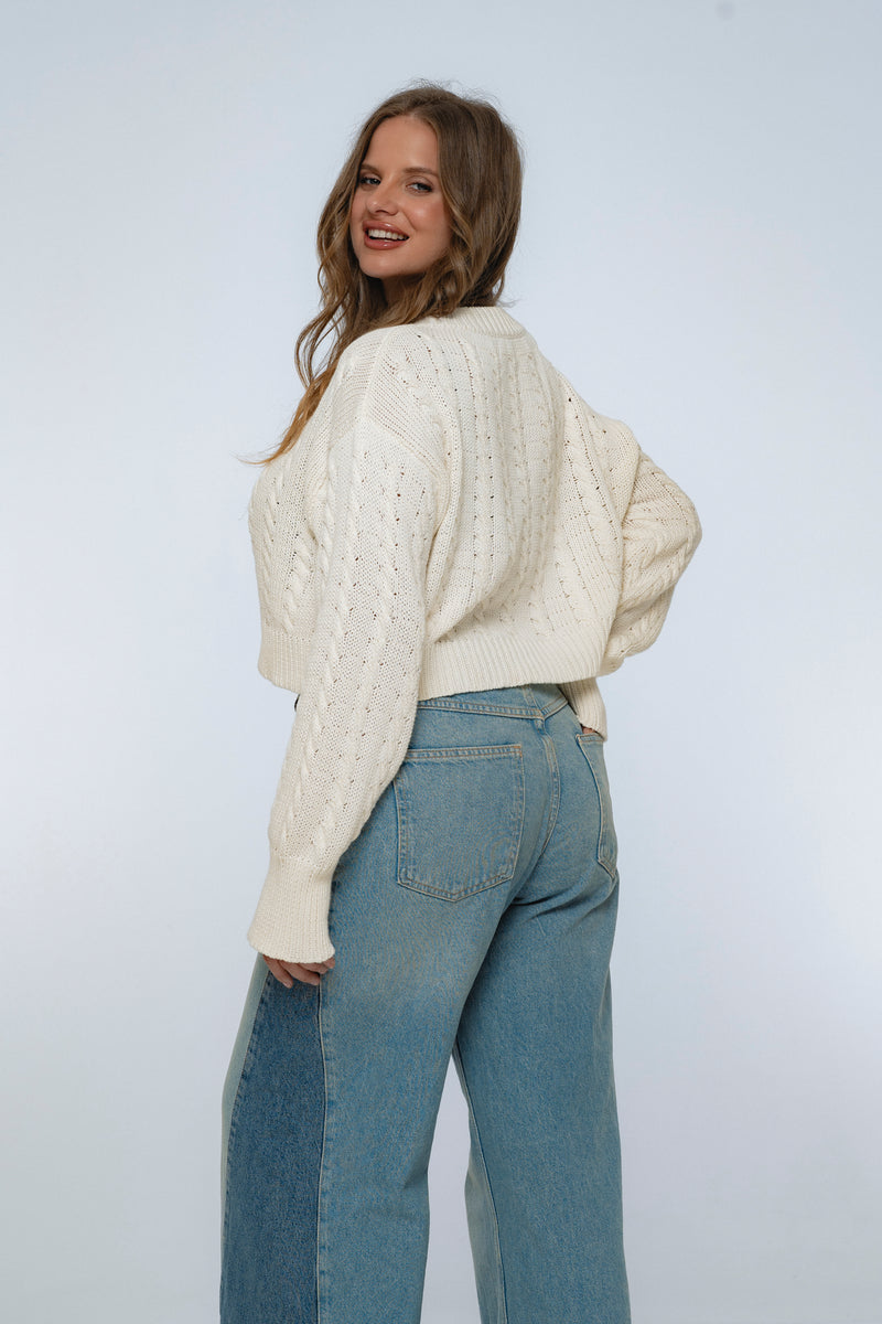 Woman wearing a cream knitted sweater and blue jeans against a white background