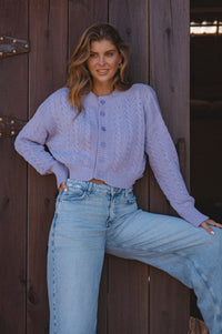Woman wearing a light purple cardigan and blue jeans leaning against a wooden door.