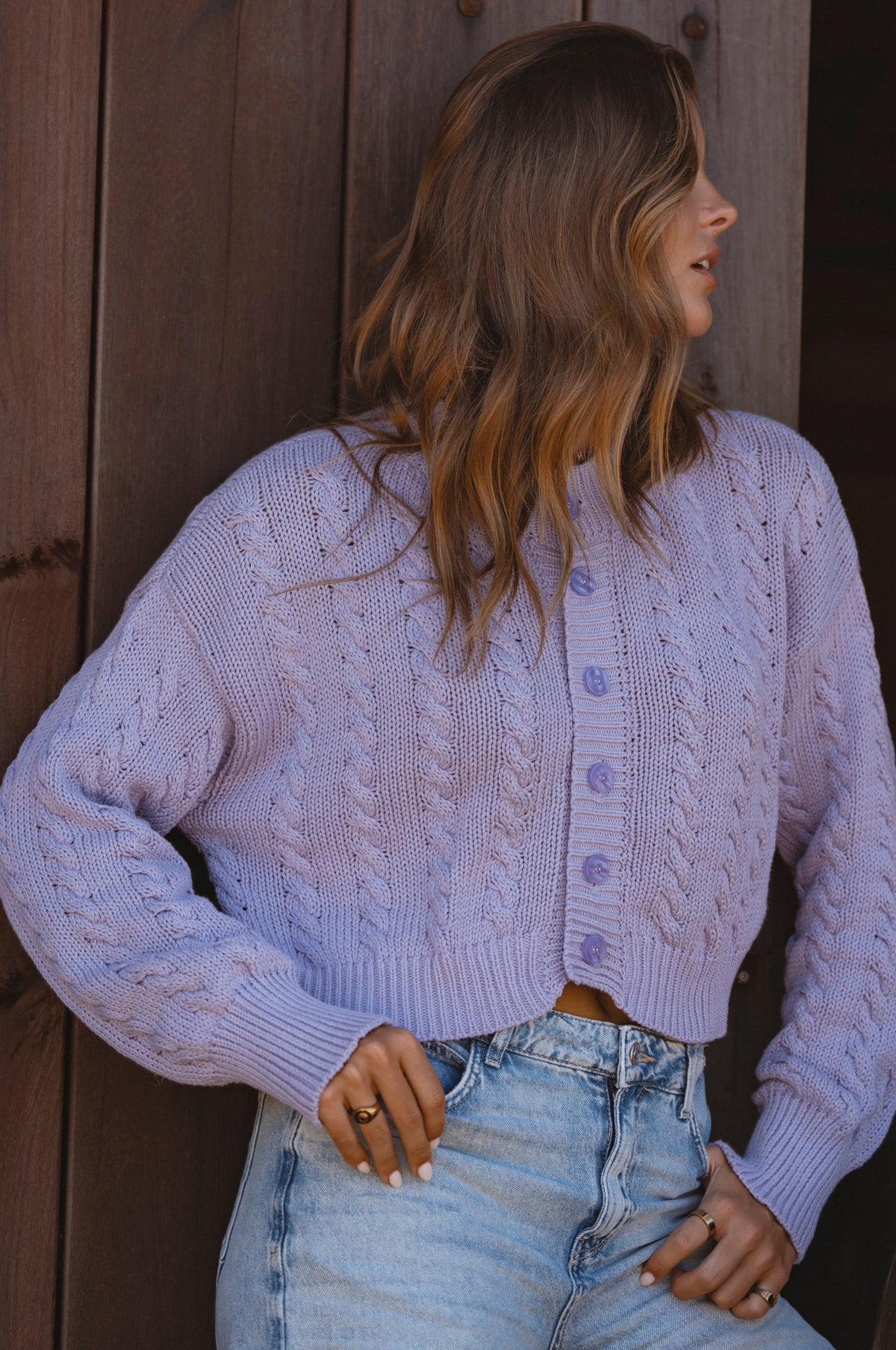 Woman wearing a light purple cable knit cardigan and denim jeans against a wooden background