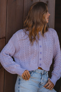 Woman wearing a light purple cable knit cardigan and denim jeans against a wooden background
