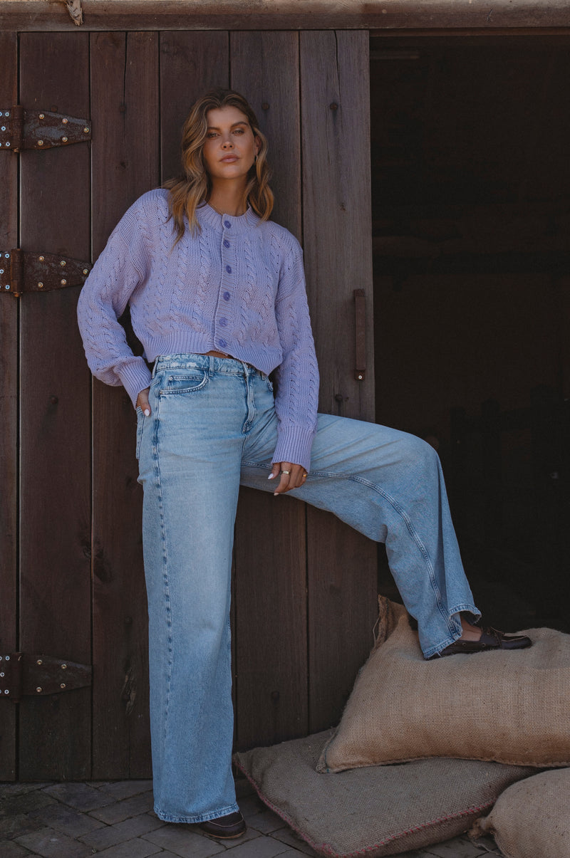 Woman wearing a light purple cardigan and blue jeans sitting against a wooden wall.