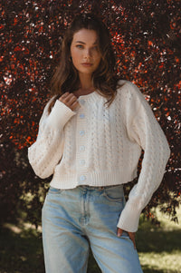 Woman wearing a white cable knit sweater and blue jeans standing against a natural background.