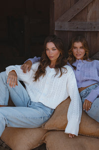 Two women sitting on a couch in a rustic setting, wearing light-colored sweaters and jeans.