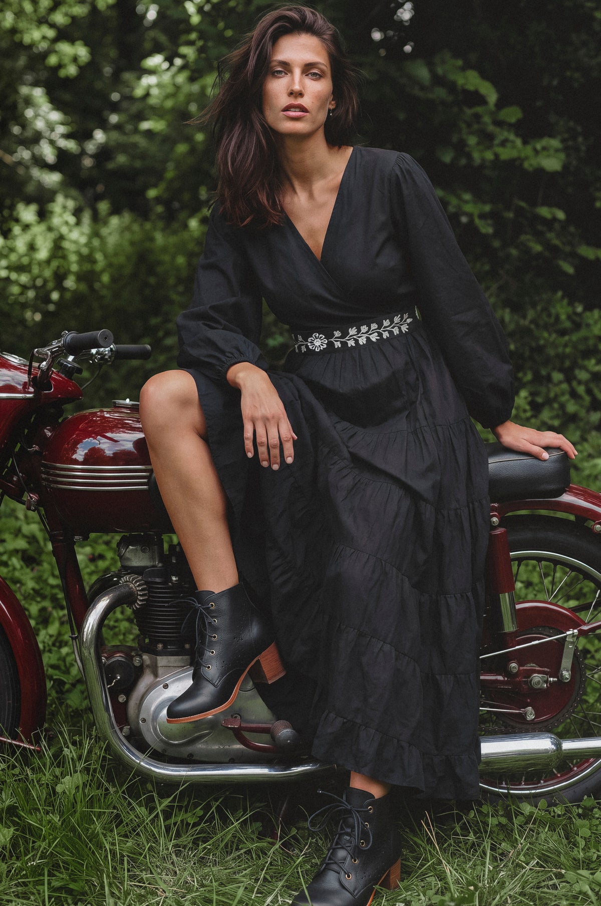 A woman wearing a long black midi dress with a silver embellished waistband and black lace-up ankle boots, sitting on a vintage red motorcycle outdoors on grass with trees in the background.