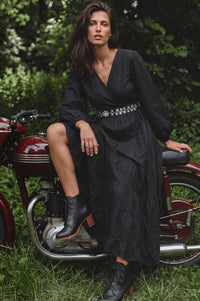 A woman wearing a long black midi dress with a silver embellished waistband and black lace-up ankle boots, sitting on a vintage red motorcycle outdoors on grass with trees in the background.