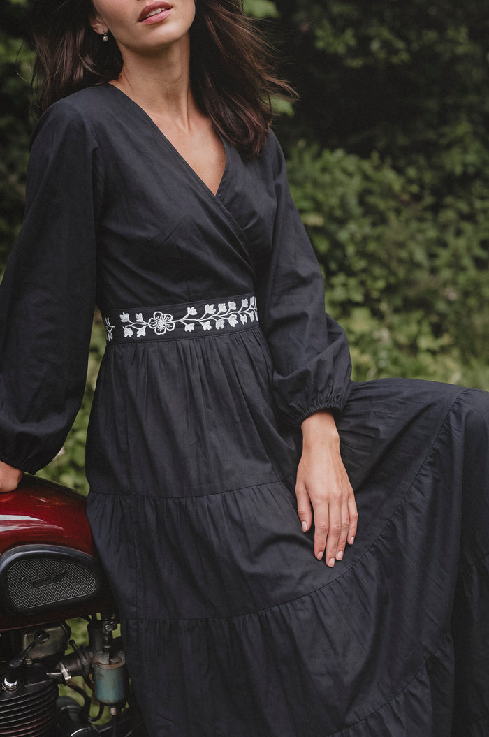 A woman wearing a black long-sleeve wrap maxi dress with white floral embroidery around the waist, leaning against a red motorcycle outdoors with green foliage in the background.