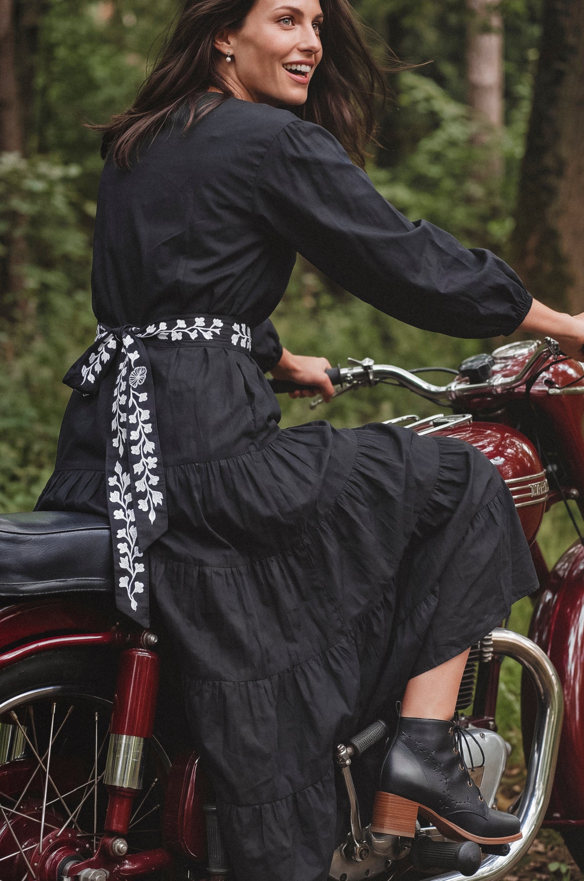 A woman wearing a long black dress with a white floral patterned waist tie sits on a vintage red motorcycle in a forest setting.