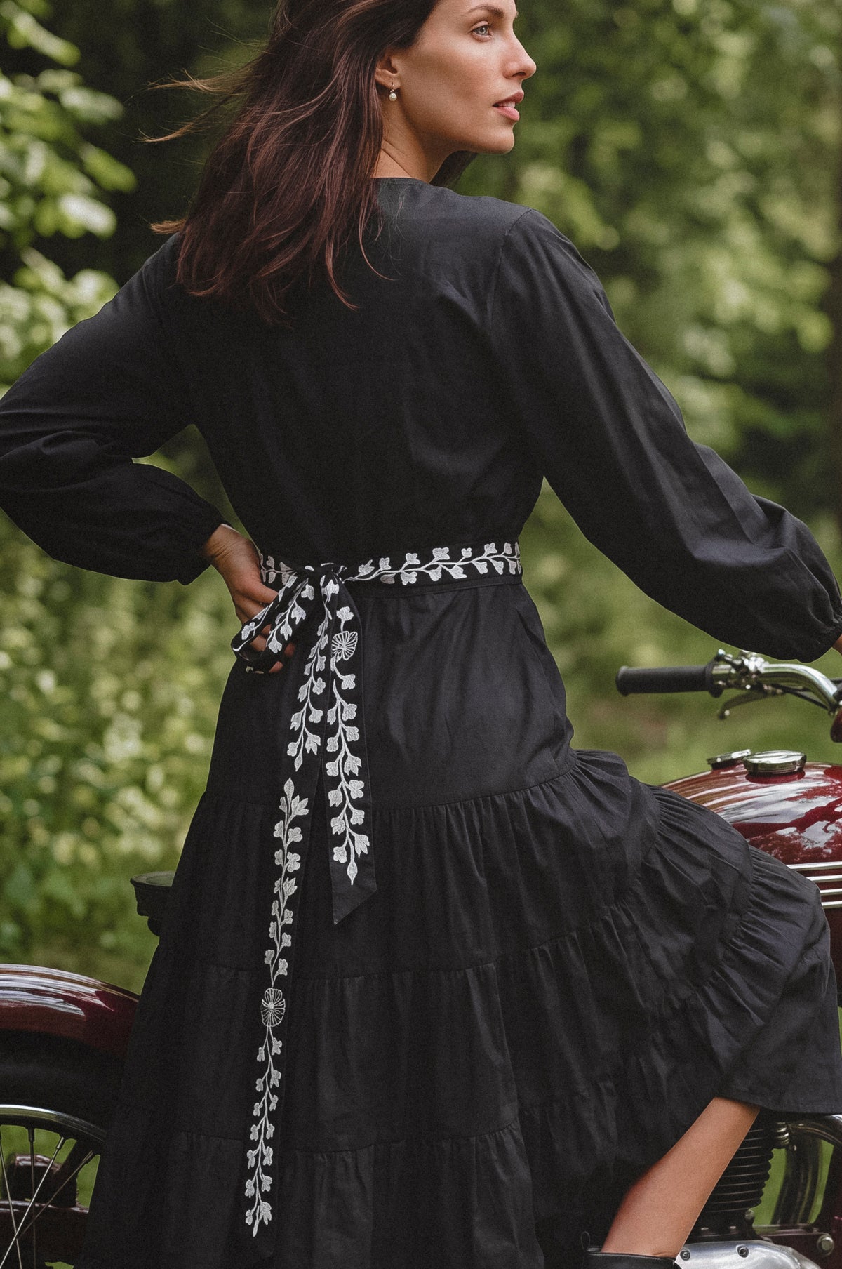 A woman wearing a black long-sleeve tiered dress with a white embroidered floral waist tie, posed outdoors on a vintage motorcycle with a blurred green forest background.