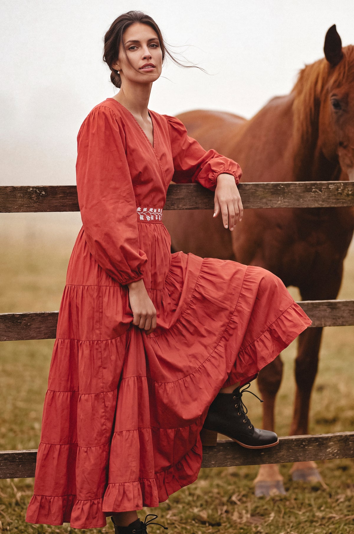 A woman wearing a rust-red tiered maxi dress with long puff sleeves and black lace-up boots leans against a wooden fence in a field, with a brown horse standing behind her.