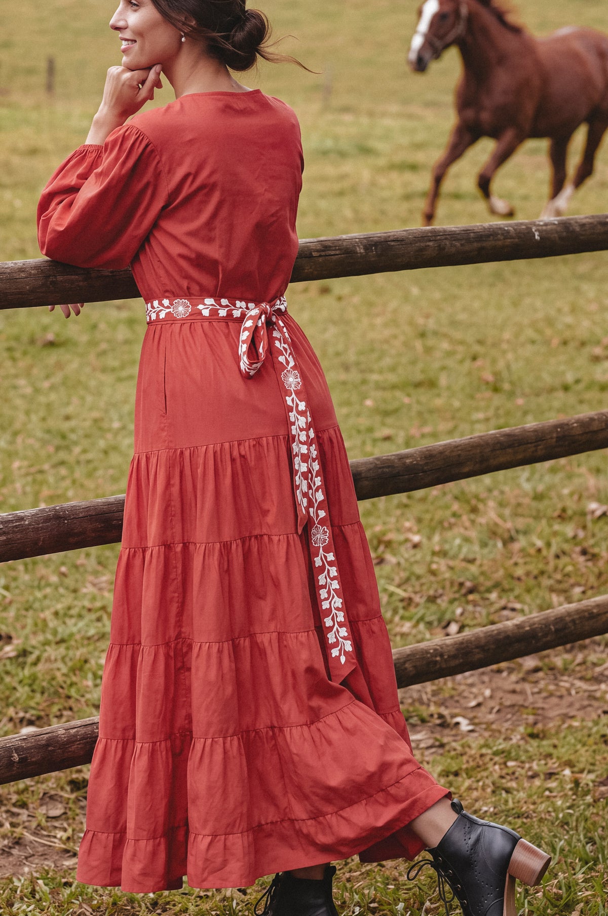 A woman wearing a rust-orange tiered maxi dress with a patterned waist tie, leaning on a wooden fence in a grassy field with a horse in the background.