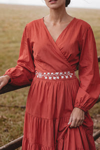 A woman wearing a rust-orange long-sleeve wrap dress with a white embroidered floral waistband, standing outdoors in a grassy field.