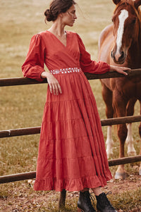 A woman wearing a rust-orange tiered midi dress with a beaded waistband leans on a wooden fence next to a brown horse in an outdoor field.