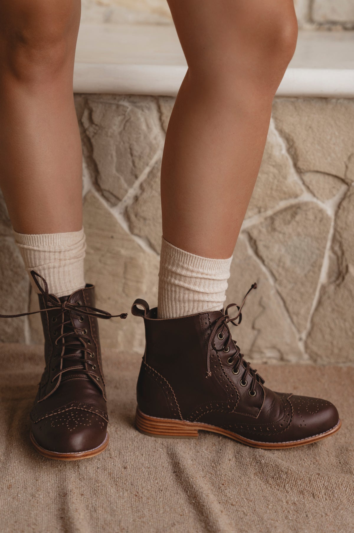 Brown lace-up boots worn with white socks against a stone wall background