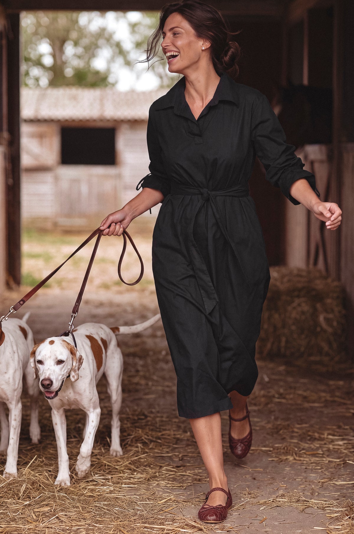 A woman wearing a black long-sleeve collared jumpsuit walks with two dogs in a barn setting with straw on the floor.