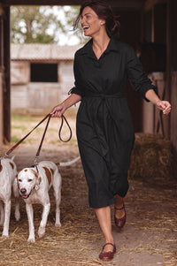 A woman wearing a black long-sleeve collared jumpsuit walks with two dogs in a barn setting with straw on the floor.