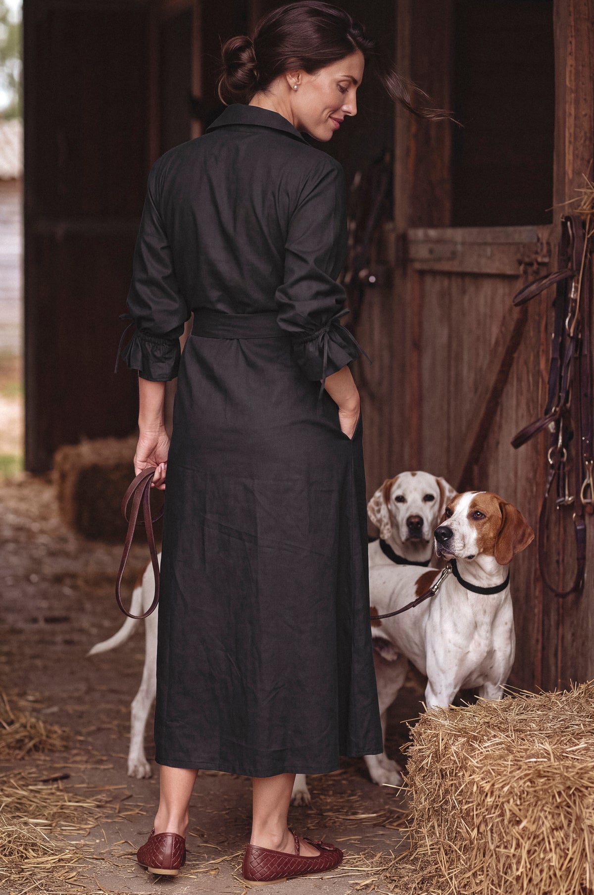 A woman wearing a black long-sleeve midi shirt dress stands in a barn with two dogs beside her. The dress has a collar, long sleeves with ruffled cuffs, a self-tie waist belt, and a straight skirt.