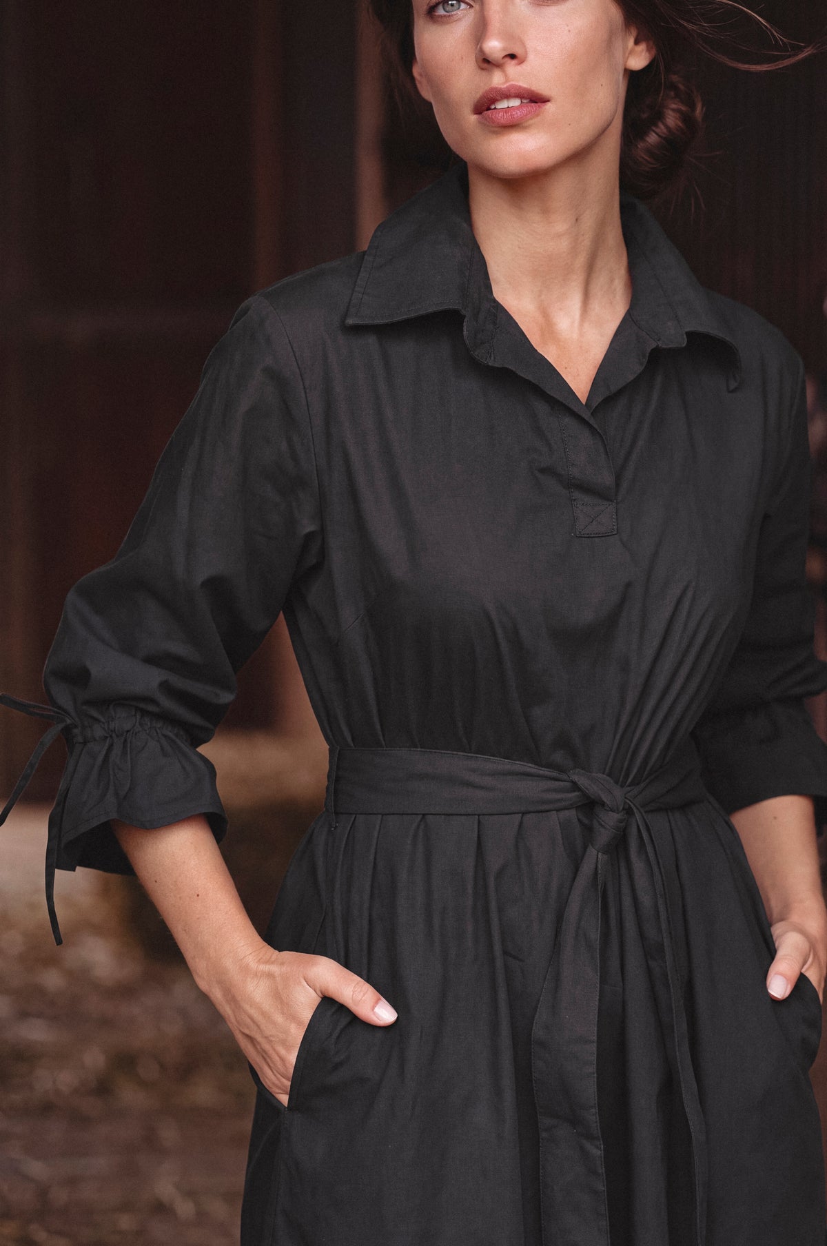 A woman wearing a black long-sleeve collared shirt dress with a tie waist belt, standing in front of a dark rustic wooden background.