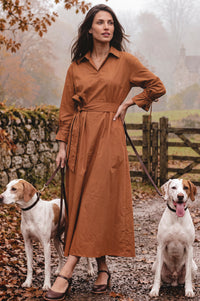 A woman wearing a rust-brown long-sleeve collared shirt dress stands outdoors on a leaf-strewn path with two dogs on leashes. The dress has a tie waist and is shown in a misty, autumnal setting.