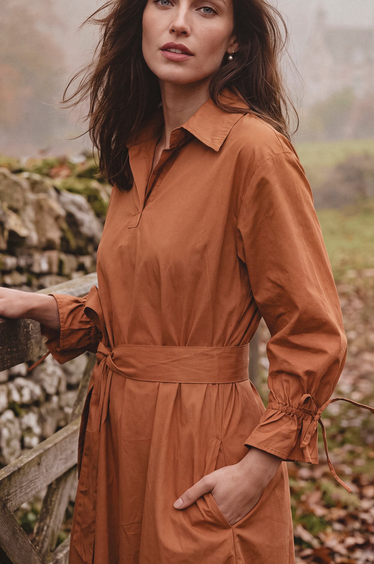 A woman wearing a rust-brown long-sleeve collared shirt dress with a matching tie waist belt, standing outdoors near a stone wall and wooden fence.