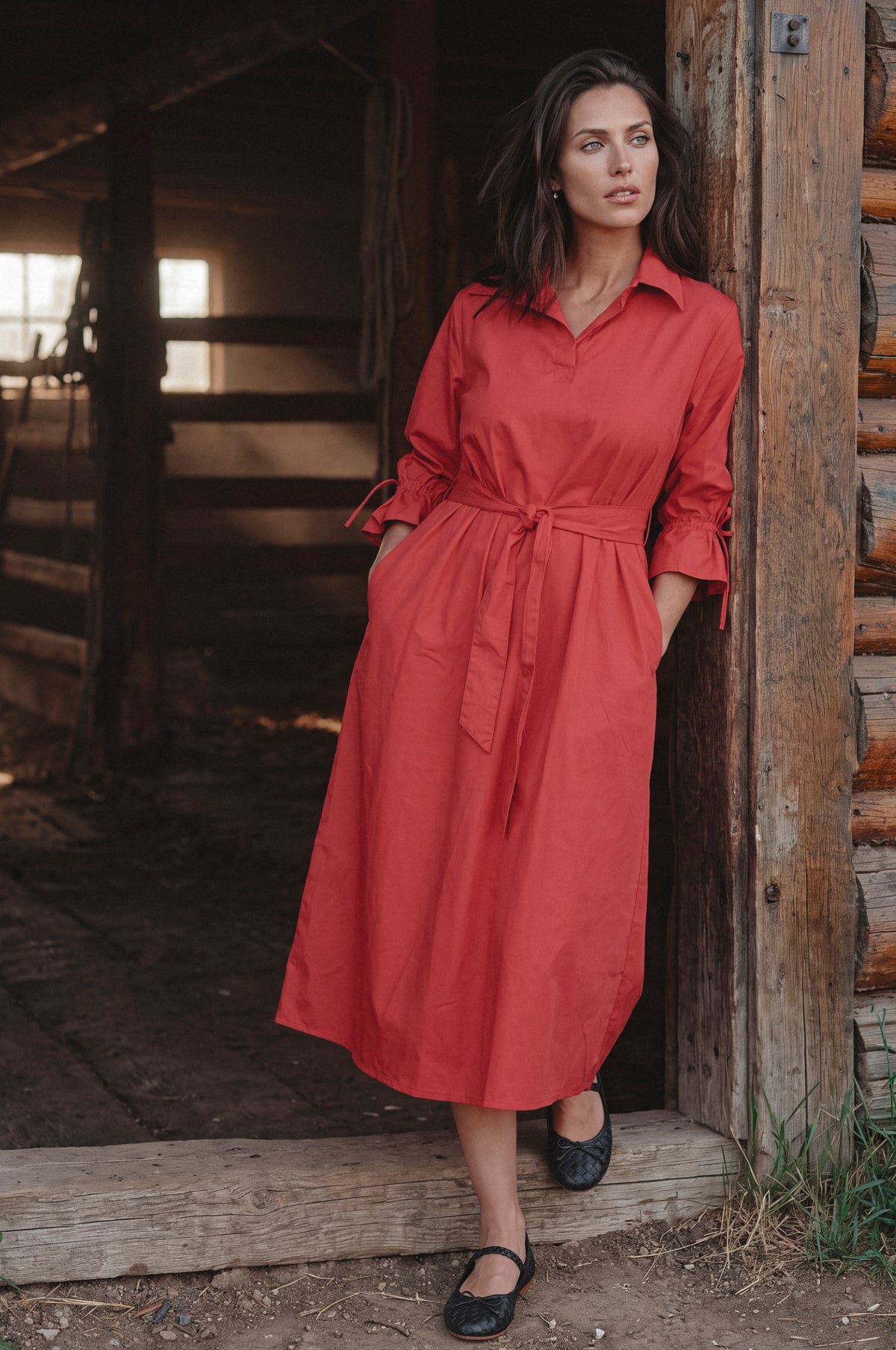 A woman wearing a rust-red midi shirt dress with a tie waist and rolled sleeves, standing in a rustic wooden doorway.